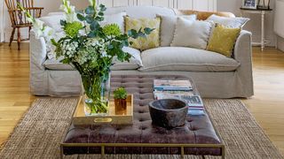 Neutral living room with wooden flooring cream linen sofa and leather buttoned stool used as a coffee table with flowers and books on display