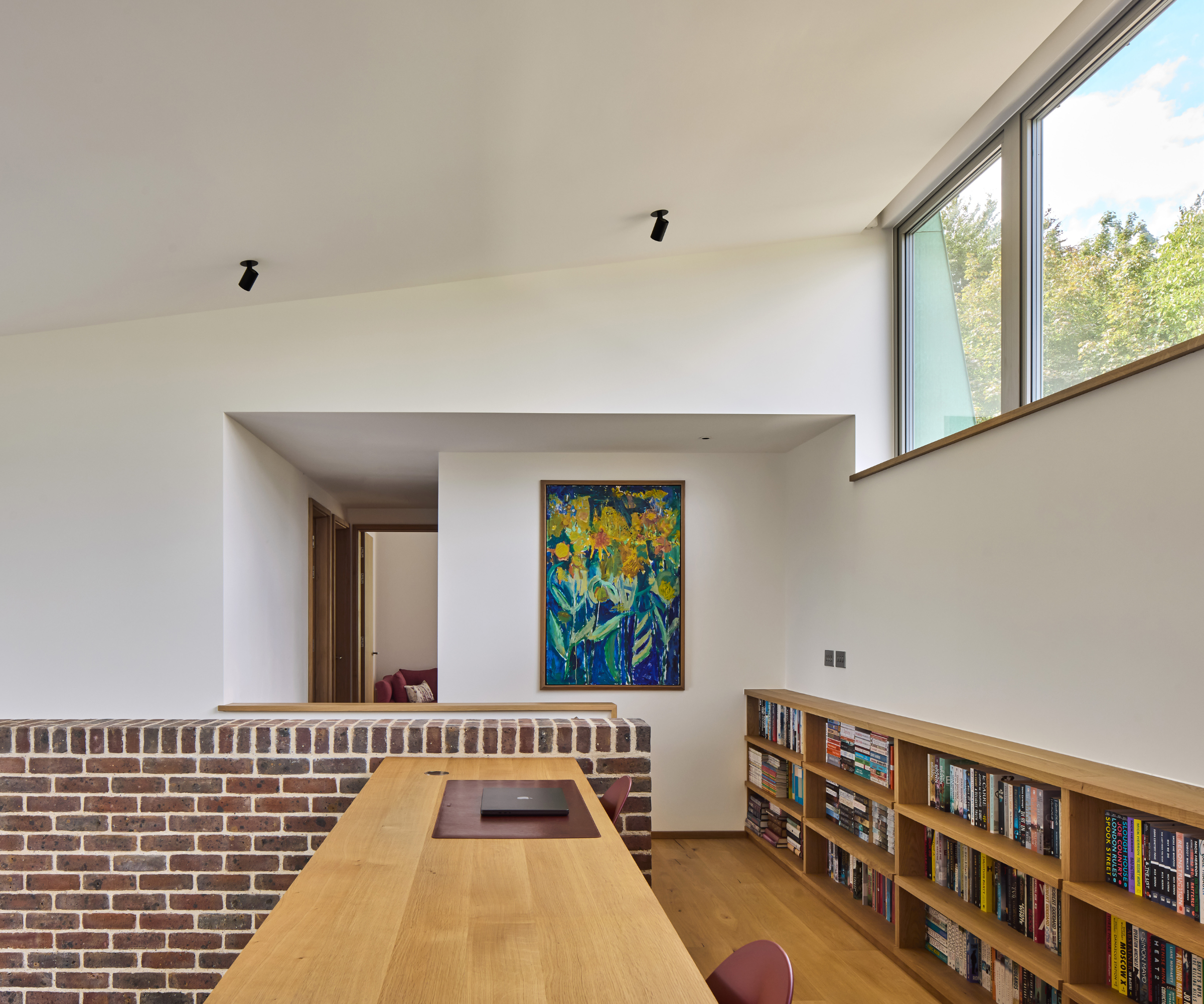 Top of the stairs case which features window and a low-level bookcase on the right hand wall, a desk on left side, and a painting straight on next to a corridor 