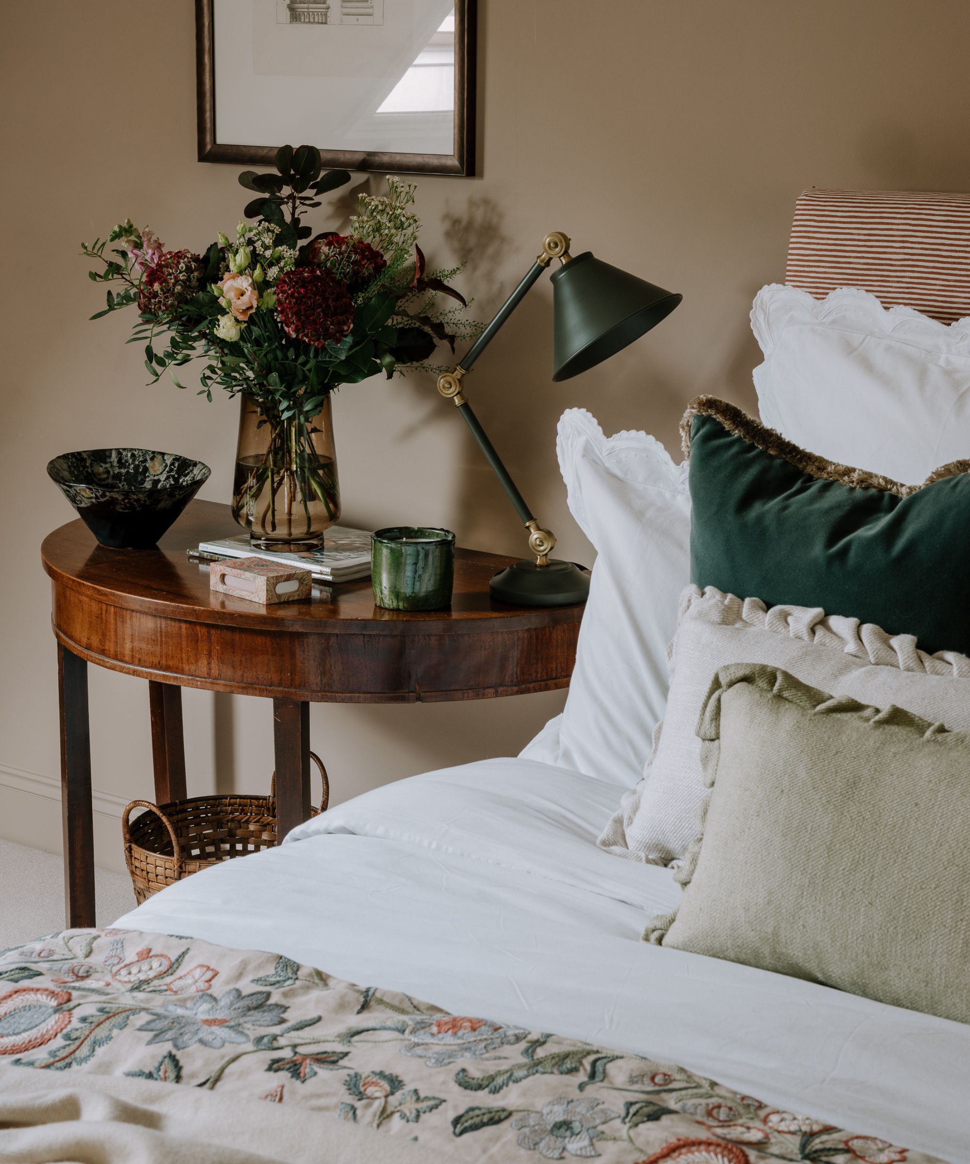 Cosy bedroom featuring a dark wood side table adorned with a green adjustable desk lamp, a vase of fresh flowers, and decorative accents. The bed is layered with plush cushions and a patterned throw