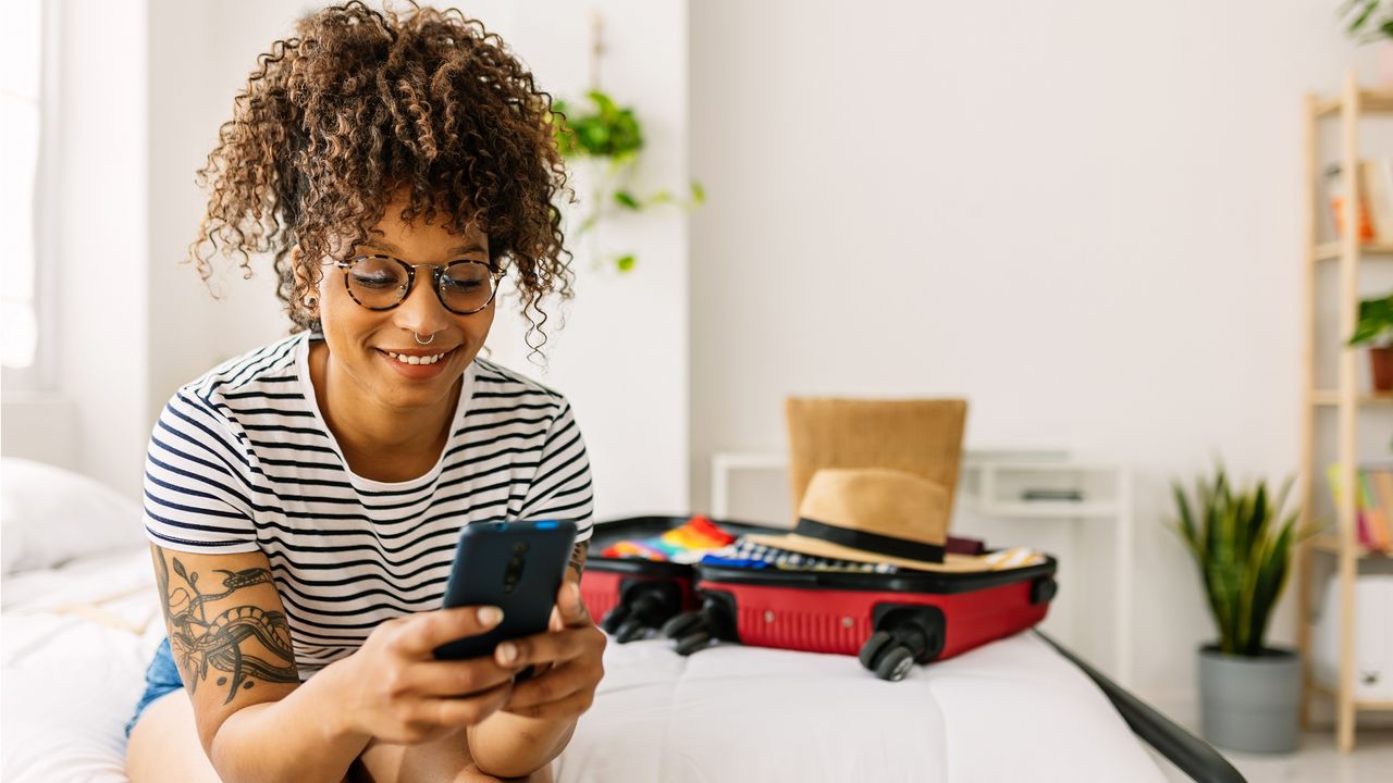 A young woman smiles while she looks at her phone in her bedroom.