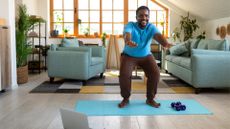 man performing a squat with arms outstretched in front on a blue exercise mat. there's a laptop on the floor in front of him and a living room setting with two sofas and a large window behind him