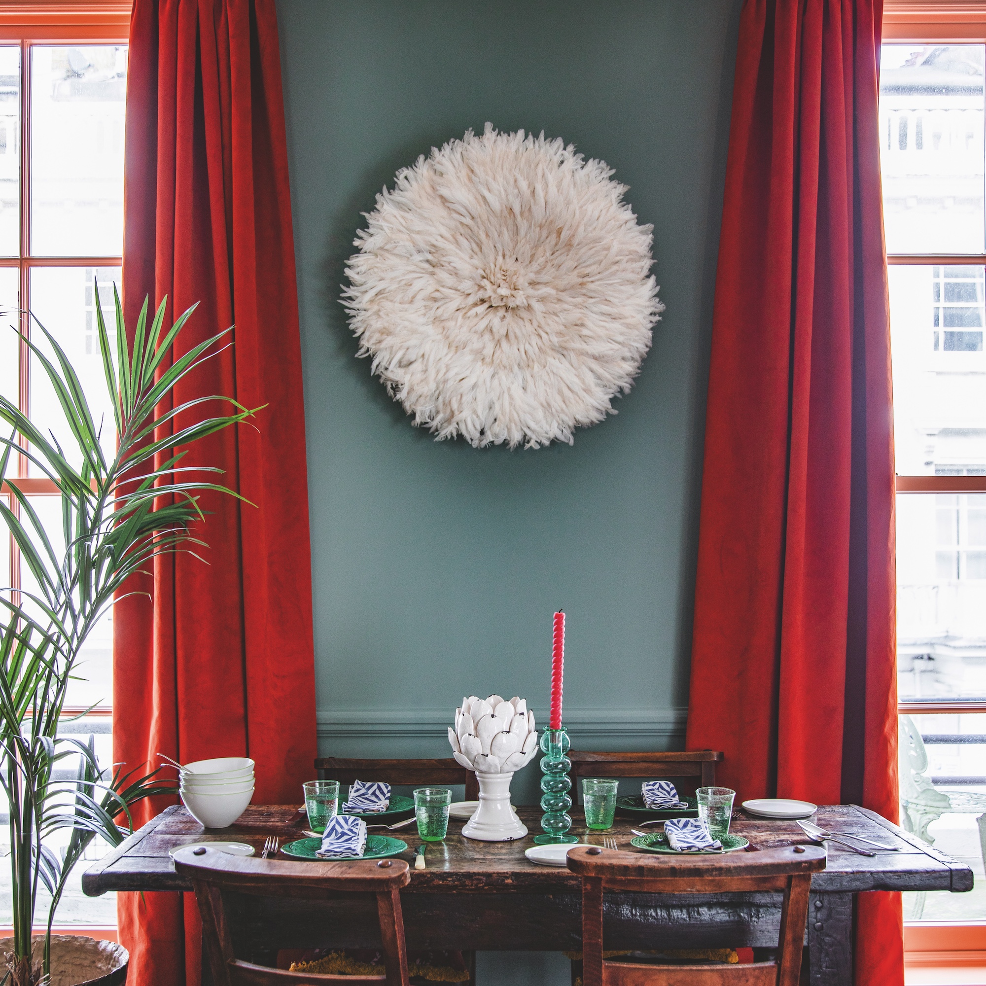 antique dining table with wooden dining chairs and mixture of green glassware and white crockery on table, in a room with blue walls and orange-red curtains