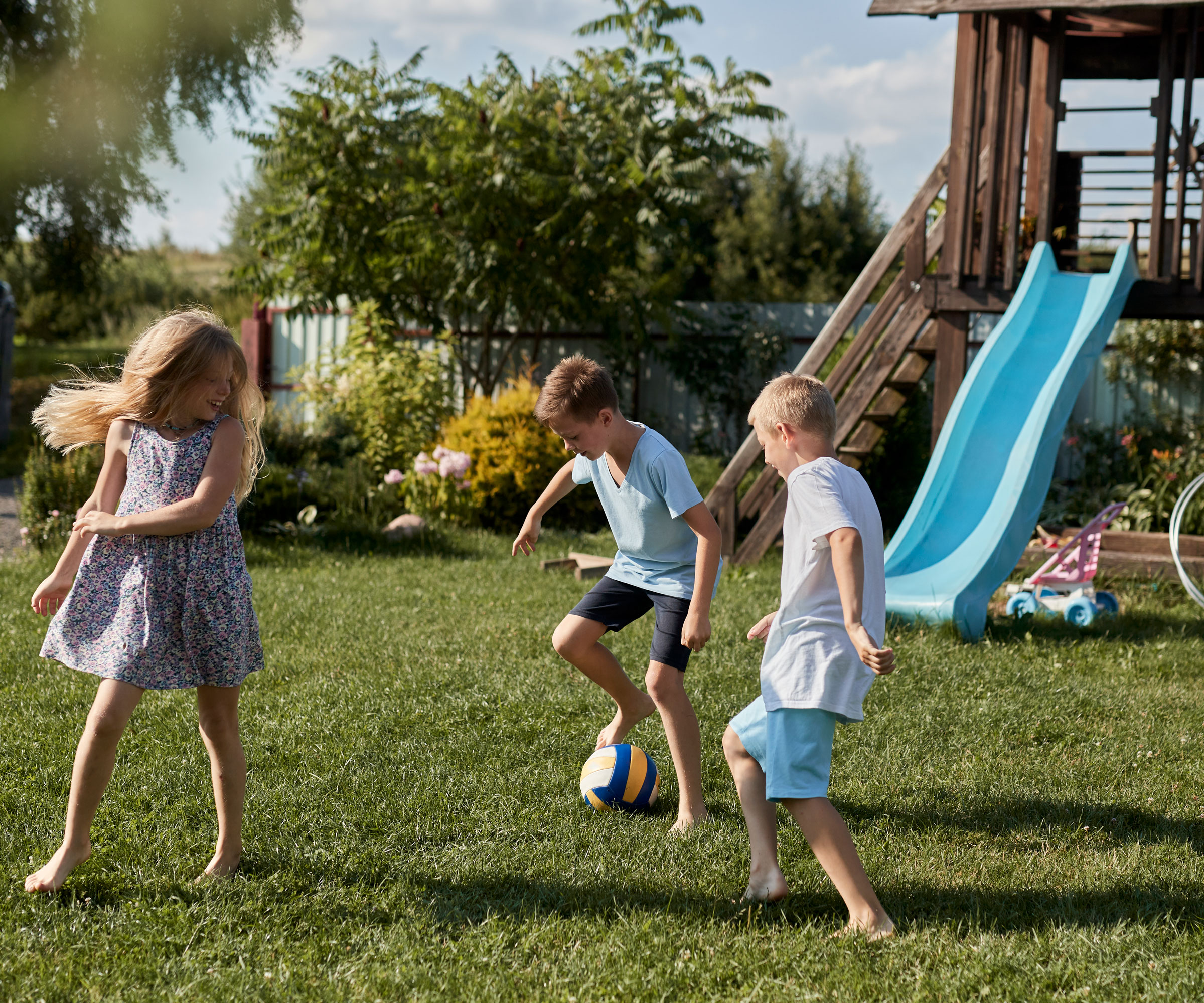 children playing football in garden with slide in the background, with trees, white fence and lawn