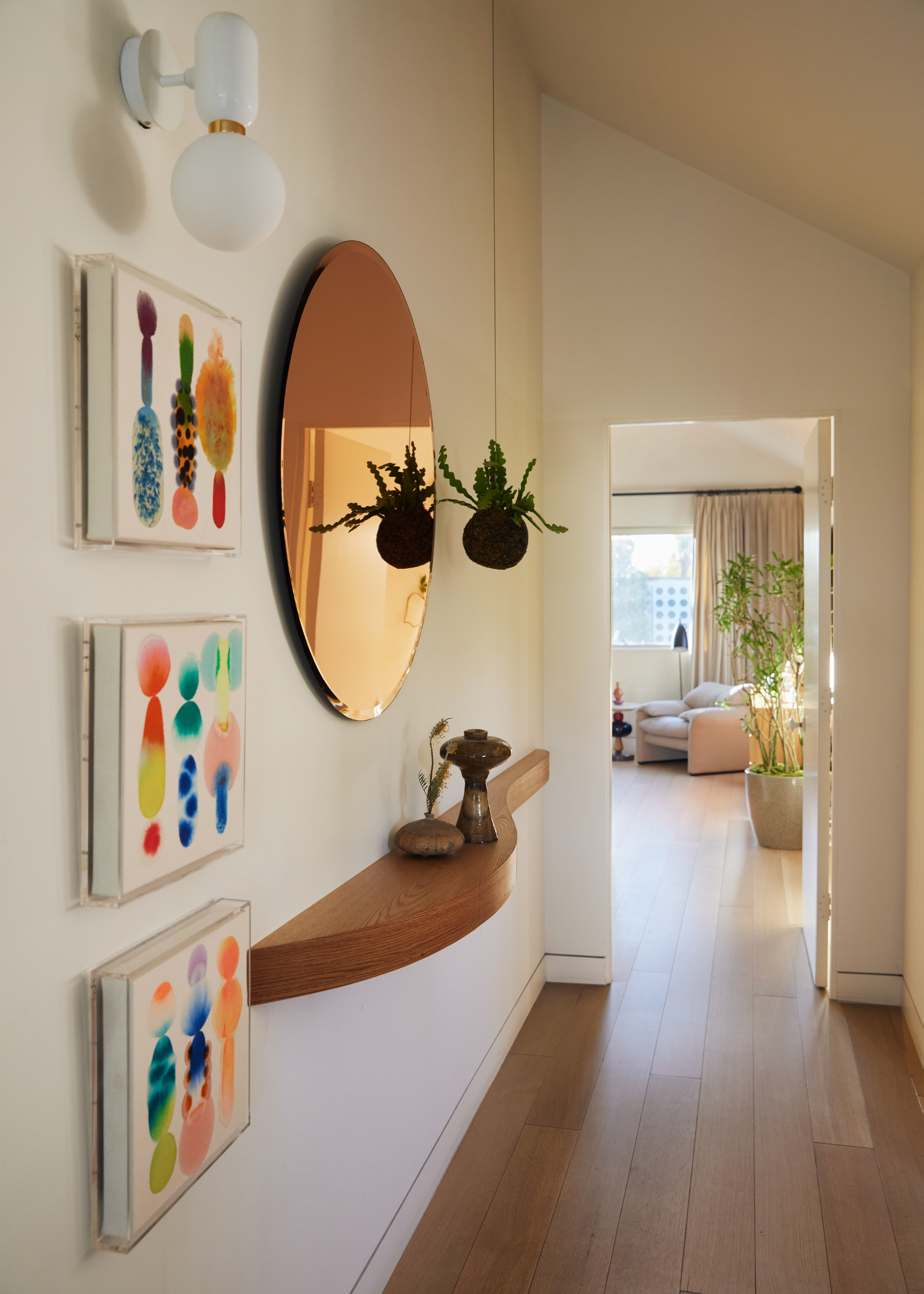 A white hallway with a floating shelf with vases below a metallic reflective circular wall accessory and a hanging planter, near a wall sconce and a trio of ink-y paintings
