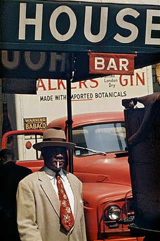 a man wearing a suit and hat with a cigarette in mouth walking in front of a red truck and shop signage