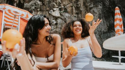 A woman holding an orange and laughing on a beach, surrounded by sun loungers and parasols.