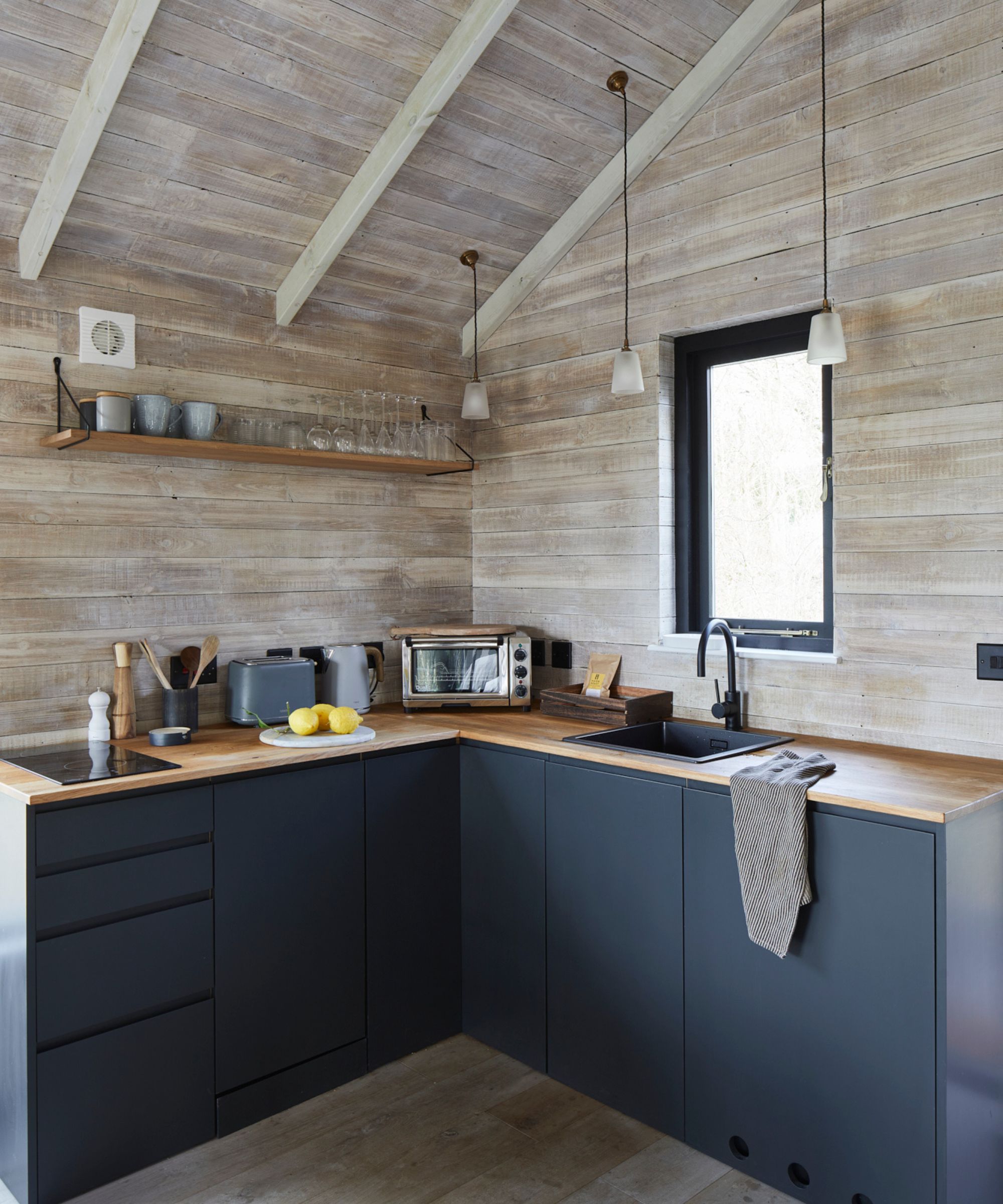 A clean and contemporary treehouse kitchen with matte navy cabinetry and a light wood countertop. Whitewashed timber cladding and exposed beams add texture, while minimalist pendant lights and a slim-framed window enhance the Scandi aesthetic.