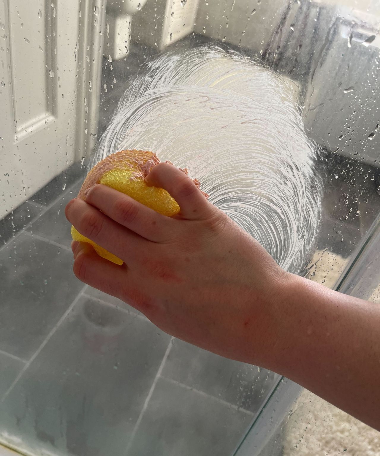 A woman's hand buffing the Pink Stuff paste onto a glass shower door with a sponge
