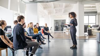 A female leader standing up to address her team, who are sat on stools to the left of the frame facing her. They are situated in a modern, brightly-lit office.