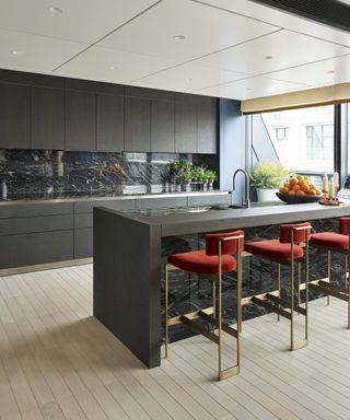 A dark kitchen with an island, light wooden flooring and red bar stools