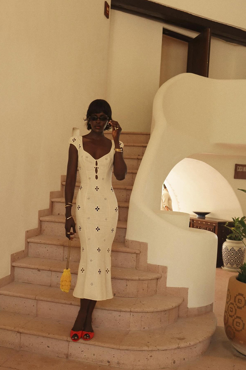 white outfit idea for summer is shown in a photo of a woman inside a hotel standing on a staircase wearing black sunglasses, gold bangles, a white midi eyelet dress, red mules, and a yellow fringe bag