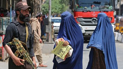 Afghan burqa-clad women walk past a Taliban security personnel