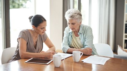 A younger woman helps an older woman with paperwork while sitting together at a table.