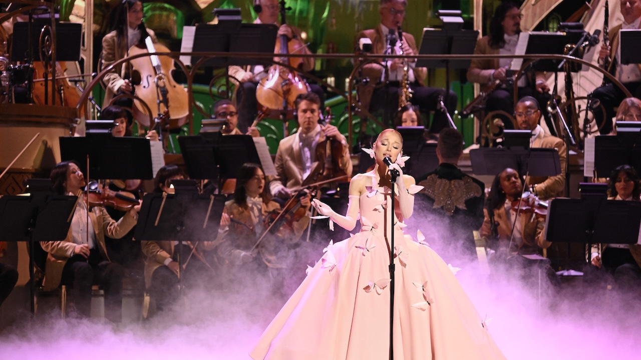 Ariana Grande in a pink ball gown singing with a full orchestra behind her