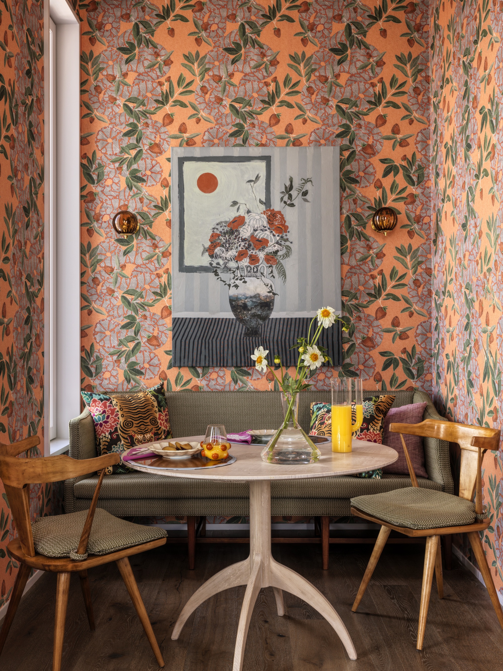 dining nook with banquette seat, round timber dining table with two dining chairs, botanical pink and orange wallpaper, with artwork, and flowers and glassware on the table
