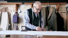 Older man with a tape measure around his neck working as a tailor, with patterns and fabric pieces hanging on the wall behind