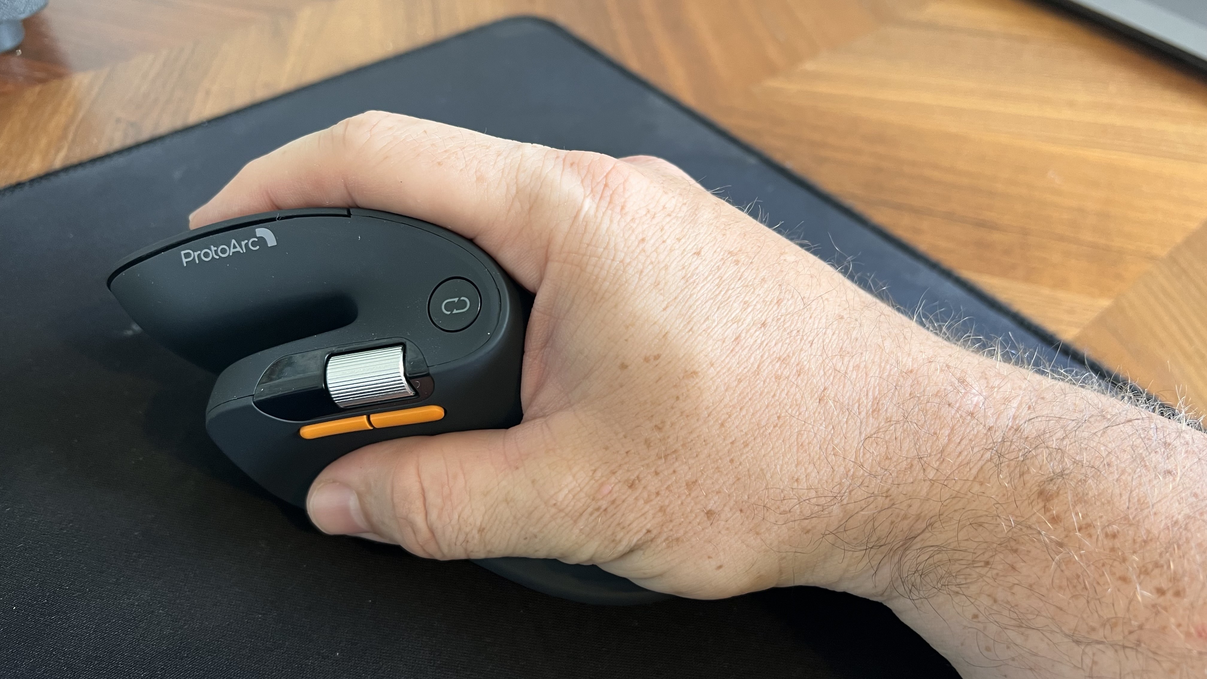 Close-up of a hand gripping the dark gray ProtoArc EM11 Pro ergonomic mouse, showcasing the thumb rest, silver scroll wheel, and vibrant orange side buttons.