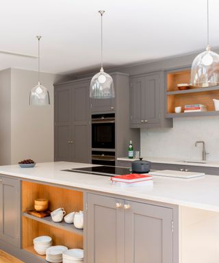 A kitchen with gray cabinets on the walls and on the kitchen island, three glass pendant lights, and a white counter with a black hob and red book on the kitchen island