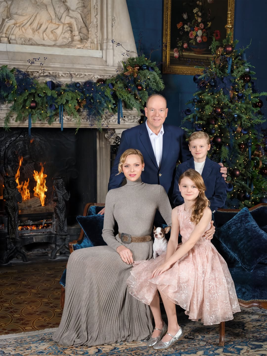 Princess Charlene wears a long taupe dress with a pleated skirt, long sleeves, and a turtle neck, and poses next to a burning fireplace and a Christmas tree with her pet dog, her twin children, Princess Gabriella and Prince Jacques, and husband Prince Albert