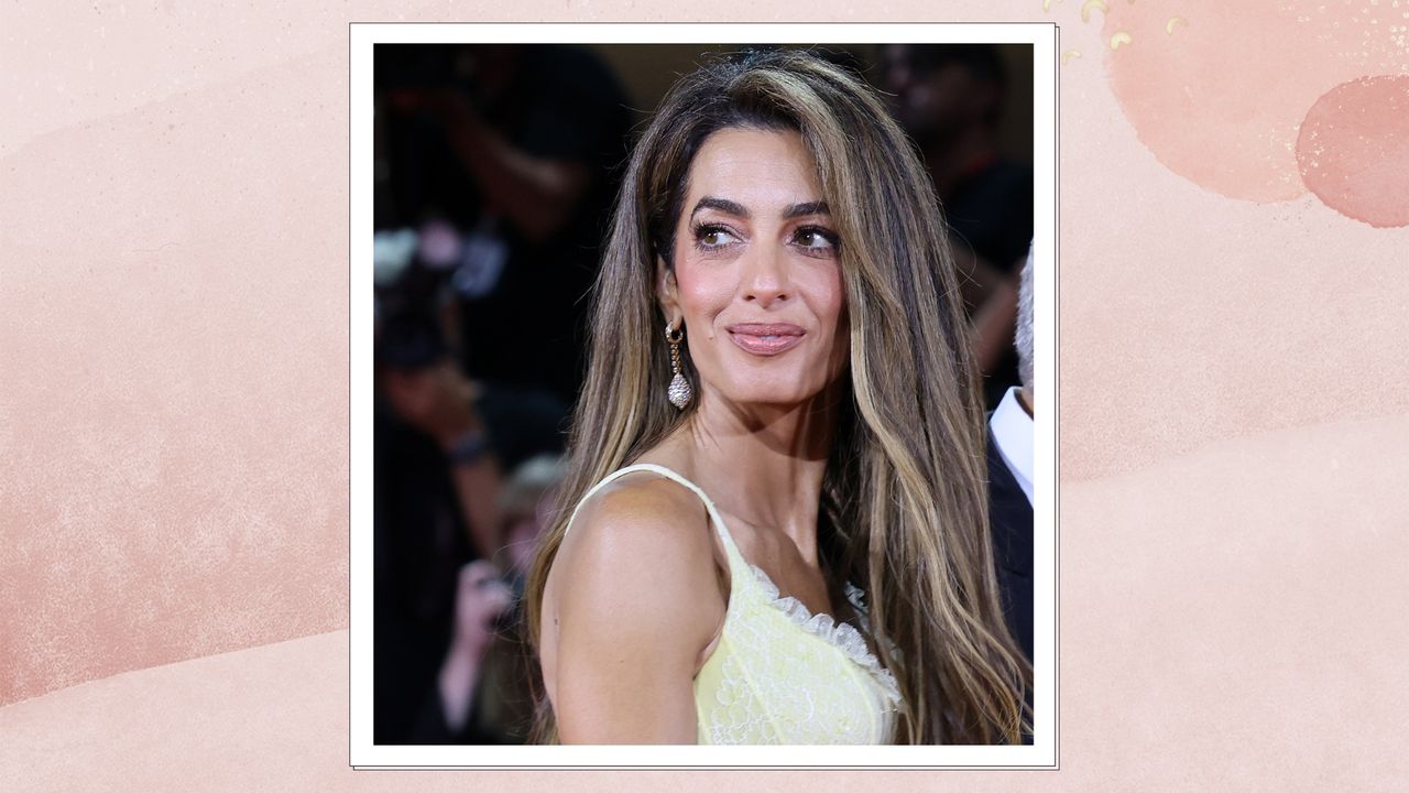 Portrait of Amal Clooney with voluminous brunette hair and blonde highlights, wearing a pastel yellow gown, on a pink watercolour background