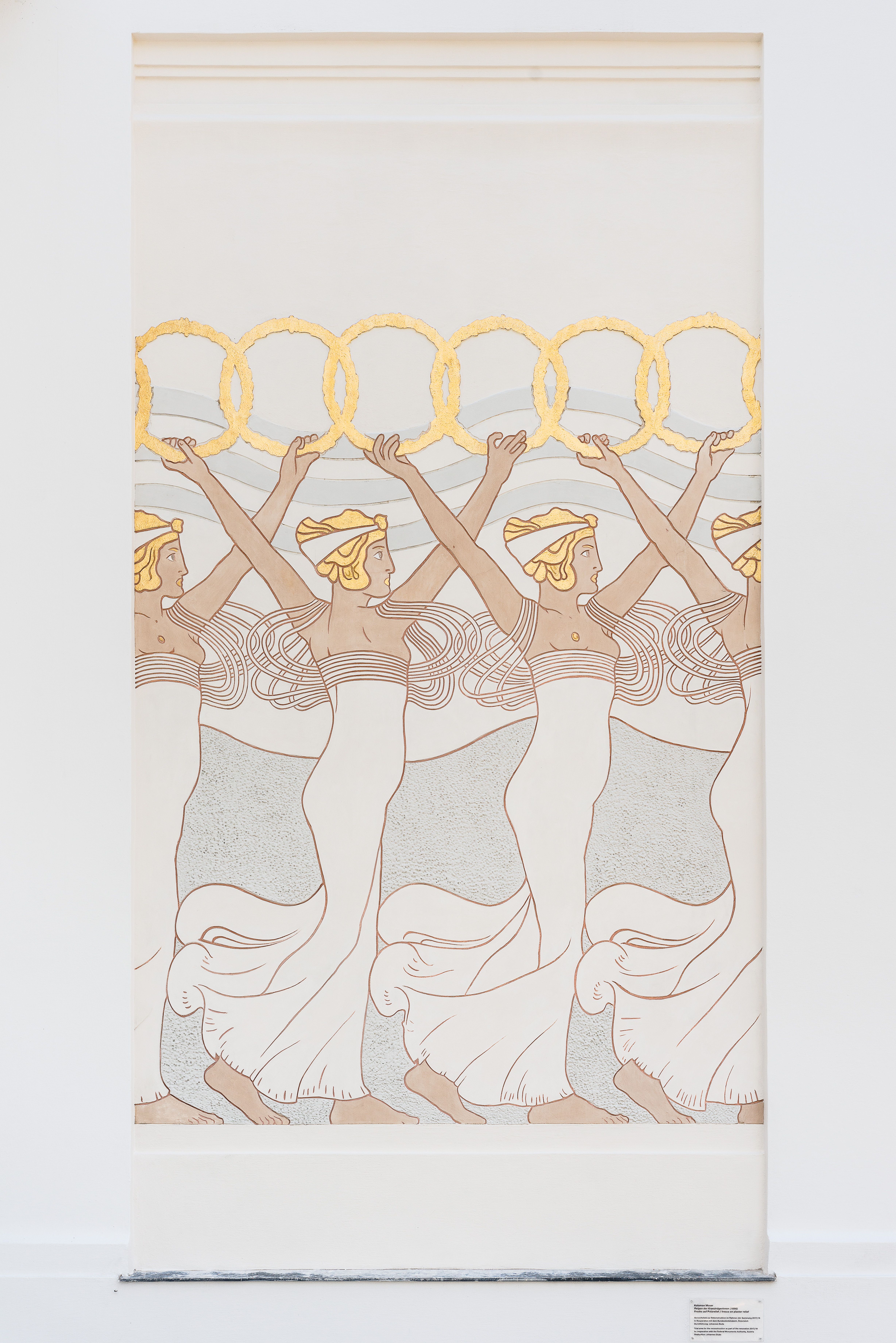 An historical building&#039;s golden-leaf frieze depicts a human procession characterized by a fantastical atmosphere and repeated floral patterns, while the rest of the facade is white.