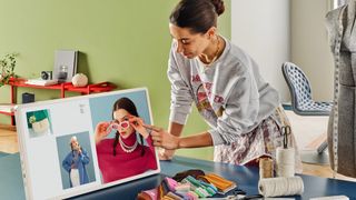 A lifestyle photo showing a woman in a bright room with a desk covered in various fabrics, scissors and threads. She is browsing fashion photos on the LG StanByME 2 TV on the desk at an angle that will give her a sore neck.