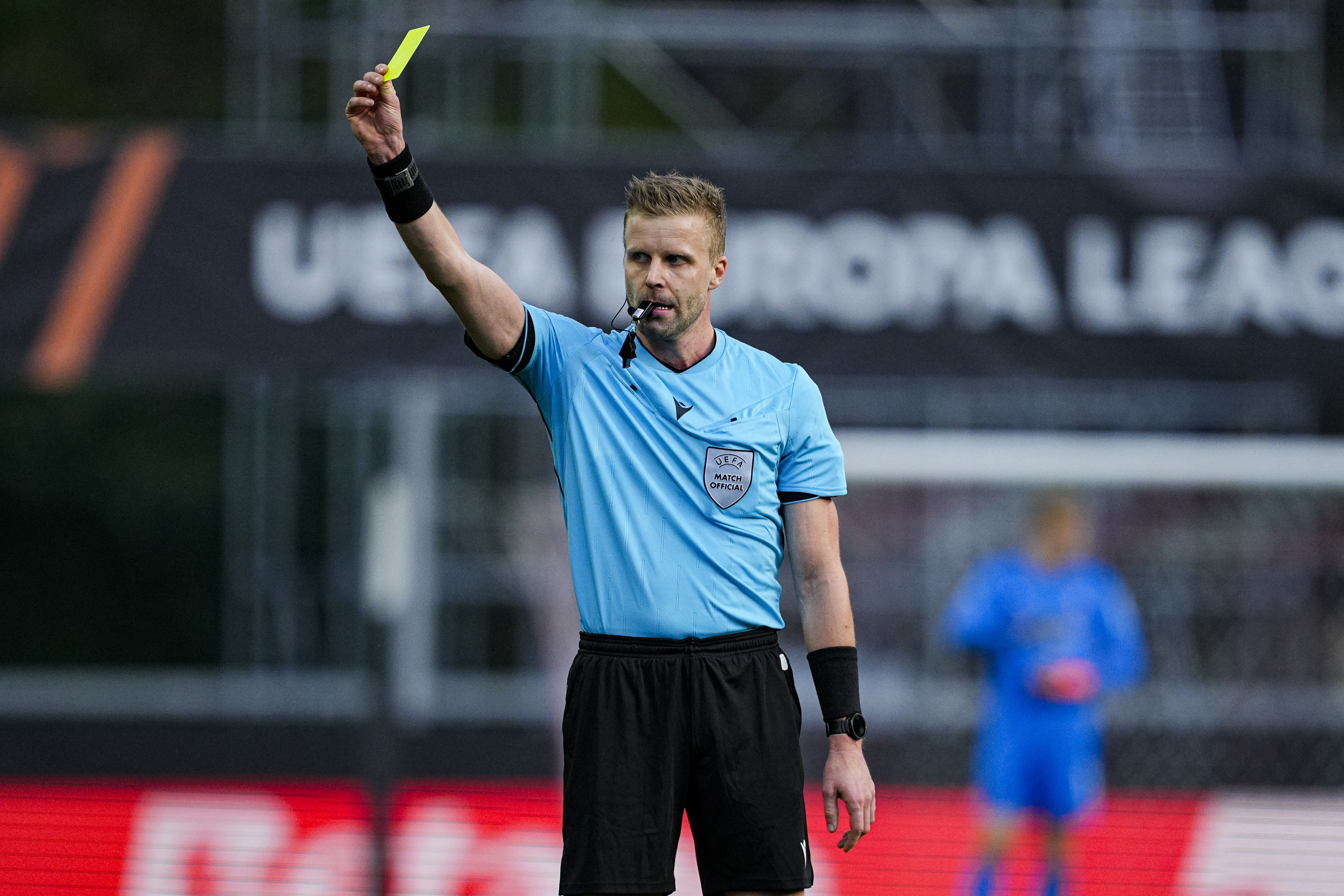 Glenn Nyberg with a whistle in his mouth, holding a yellow card above his head.