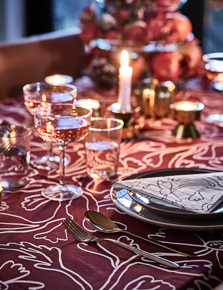 Richly layered fall tablescape featuring botanical-motif oxblood table linens, faceted champagne coupes, and brass candlesticks. 