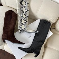 A photo of three pairs of Khaite knee-high boots on a cream-colored couch next to a Khaite boot box.