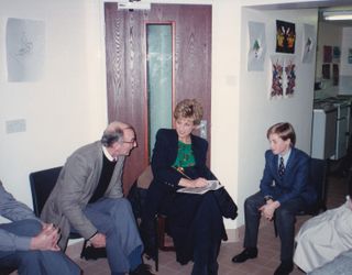 Princess Diana sitting in a chair talking to a man while Prince William sits next to her at a homeless shelter