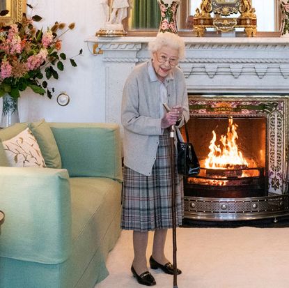 Queen Elizabeth wearing a plaid skirt and gray cardigan leaning on a cane and standing in front of a fireplace