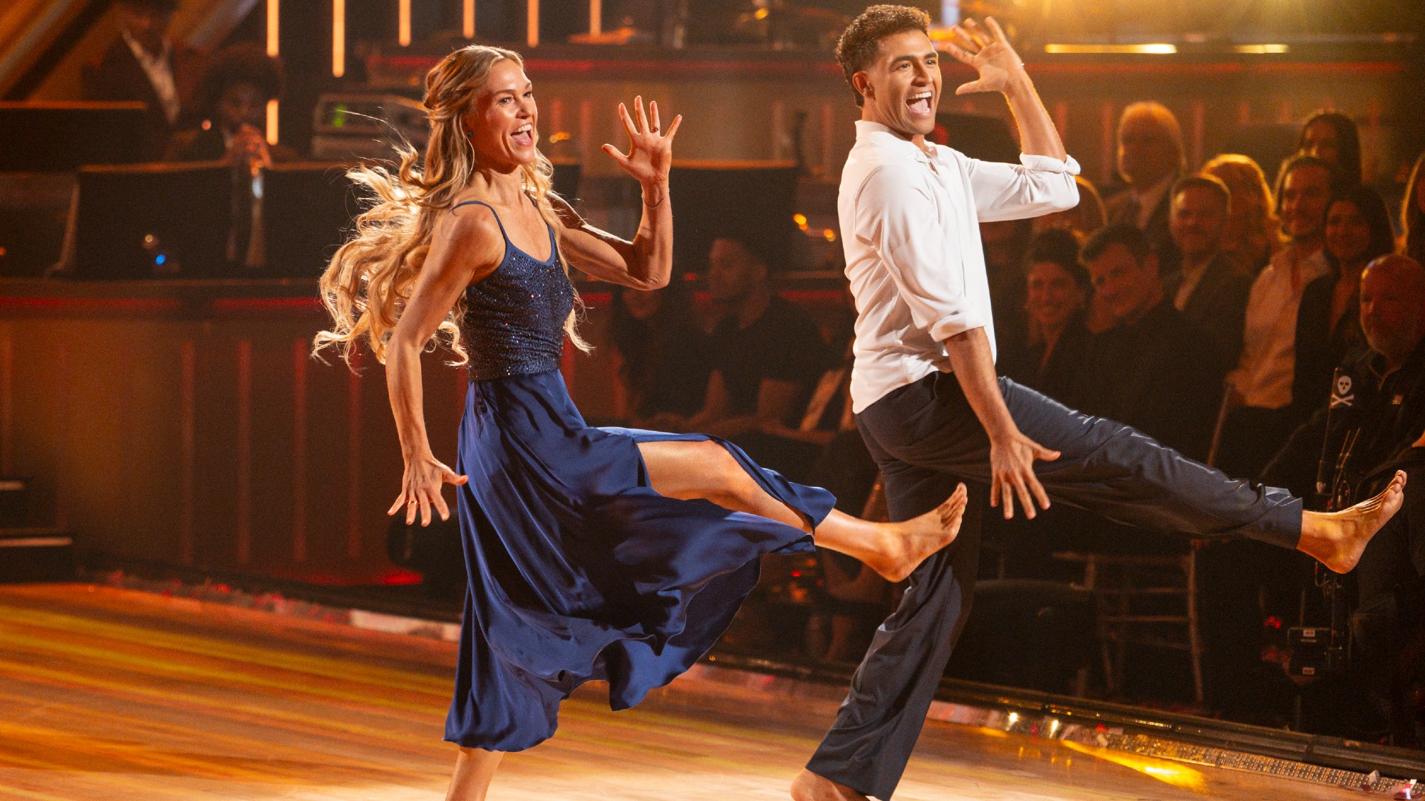 Julia and Ezra doing a leg kick on Dedication Night. Julia is wearing a blue dress. Ezra is wearing a white shirt and black slacks. 