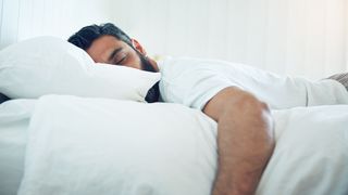 A man with dark hair wearing a white t-shirt sleeps on his stomach on a white bed