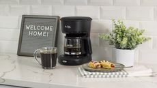 Mr. Coffee 5-Cup Mini Brew Coffee Maker on marble countertop with signage, pastries, dish towel and indoor houseplant