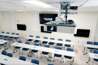 Active learning classrooms have a flexible setup and can be reconfigured to address semester-based pedagogical needs. An Extron NAV E 101 is installed with the PTZ camera over the suspended exam table to send images to the Distance Education Control Room over the UNCW network. This classroom is shown in the traditional configuration, which is better for social distancing.