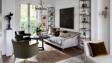 A bright living room with white walls, wooden flooring, a velvet sofa, green armchair, white sofa with velvet cushion, tall shelving, circular coffee table and a large chandelier. Above the sofa is a statement piece of dark artwork. Behind are french doors to a bright and leafy garden.
