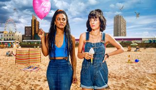 Freema Agyeman and Lily Allen in character as Trish and Mel in Dreamland. They are standing on the beach with Dreamland amusement park in the background behind them, and they are looking at each other warily. Trish is holding a 'mum to be' balloon, and Mel is holding a 99 ice cream