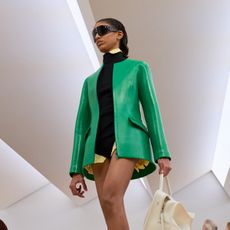A model at the SS26 Loewe show wearing a green leather sculpted blazer with a sweater and white bag.