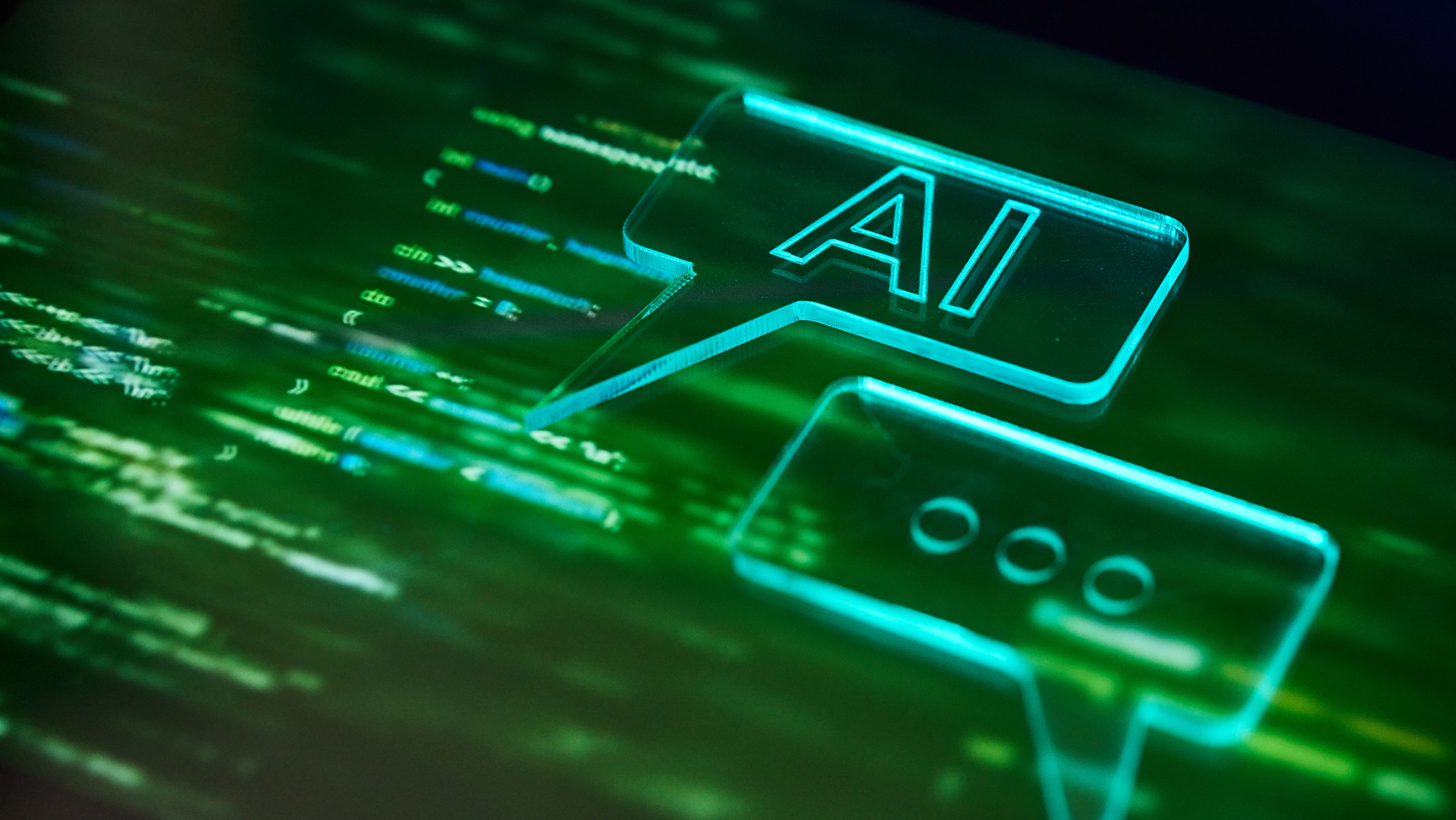 An abstract image of green code with two blue speech bubbles on top, one reading 'AI' and the other showing three dots.