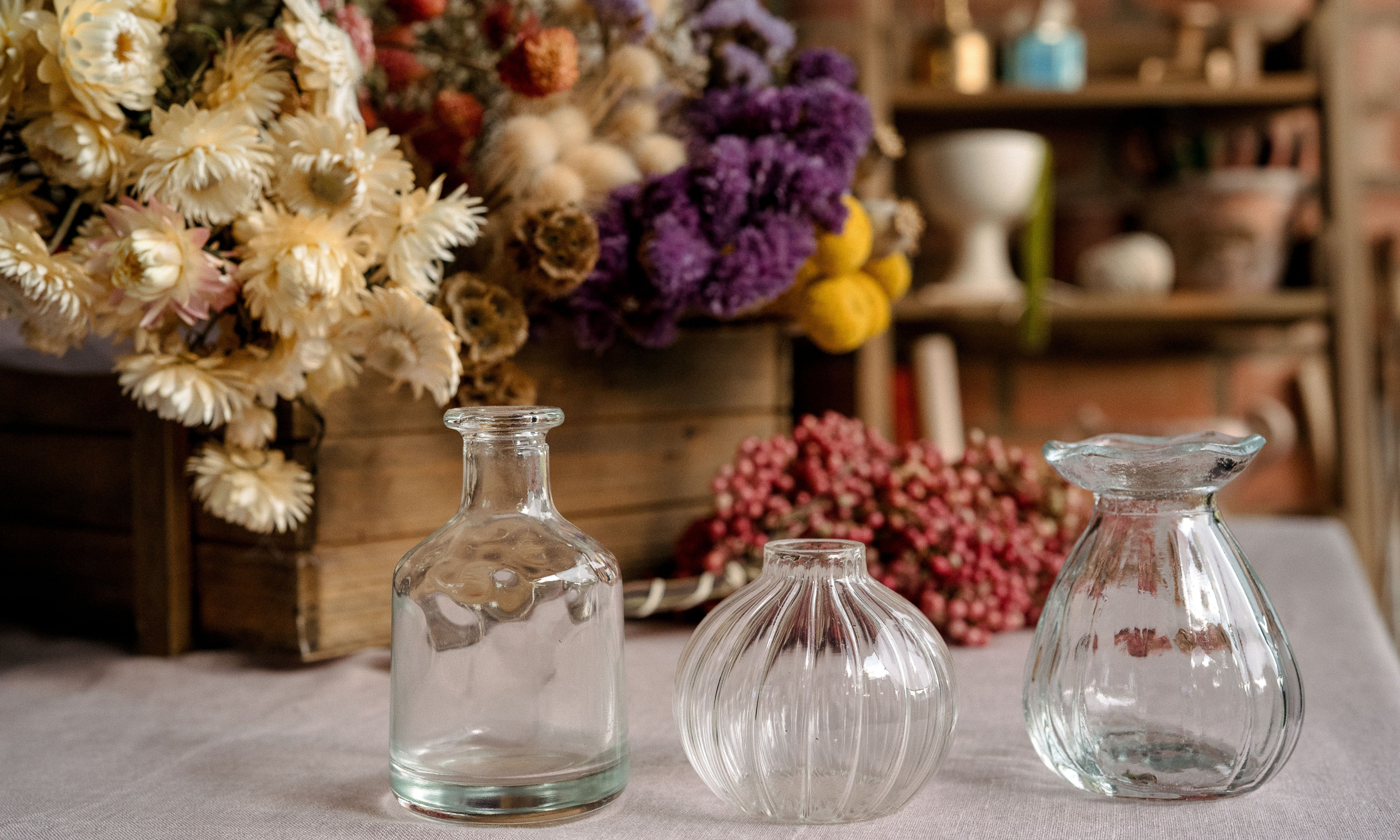 Three clear glass bud vases on a table, with bunches of dried flowers and pink peppercorns behind