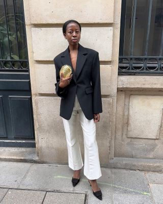 stylist and influencer Sylvie Mus poses on a Paris sidewalk wearing a black tailored blazer, small metallic clutch bag, white flared pants, and black suede mule pumps