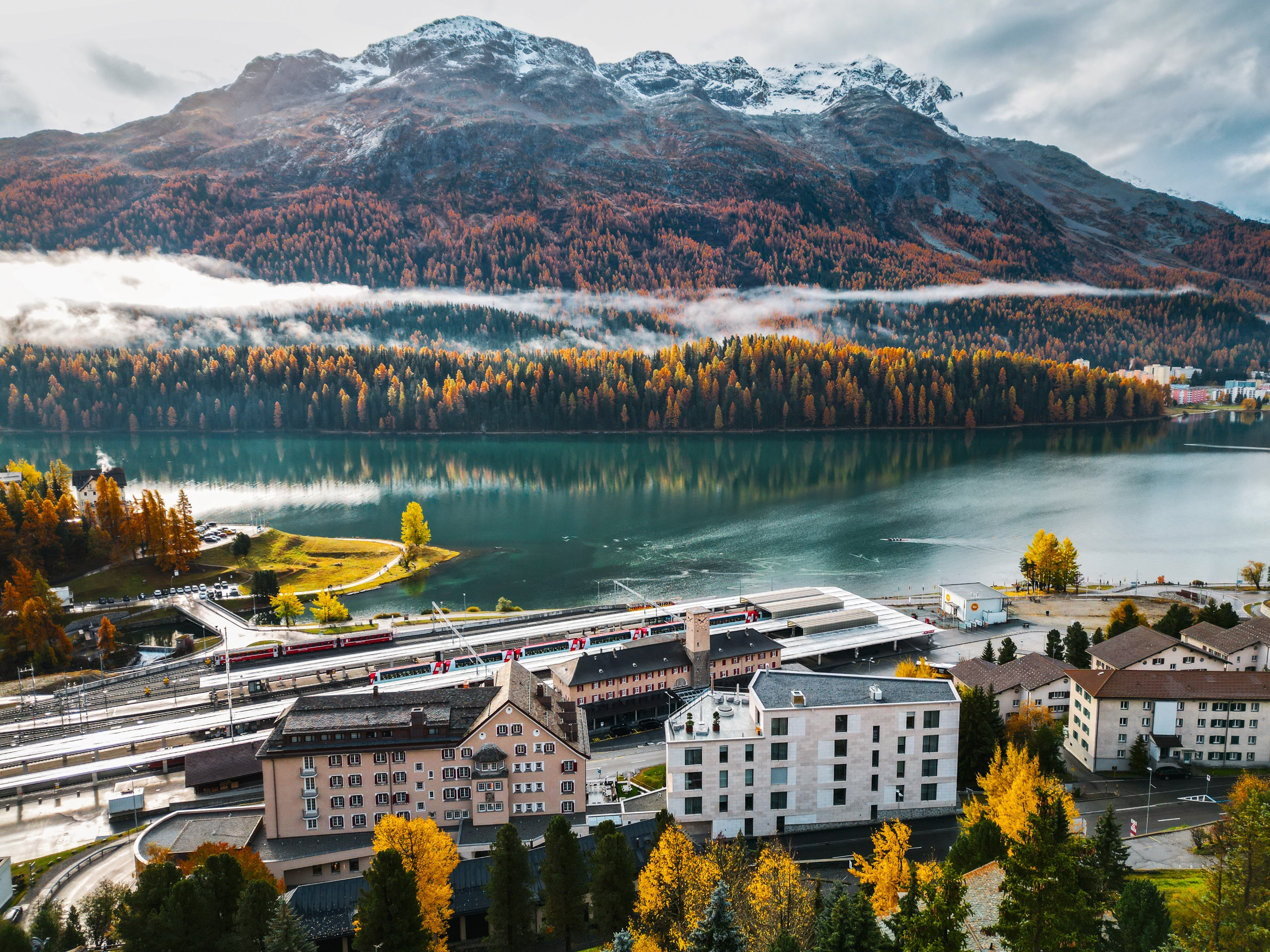 Buildings and a railway terminal infront of an alpine lake and snow-capped mountains