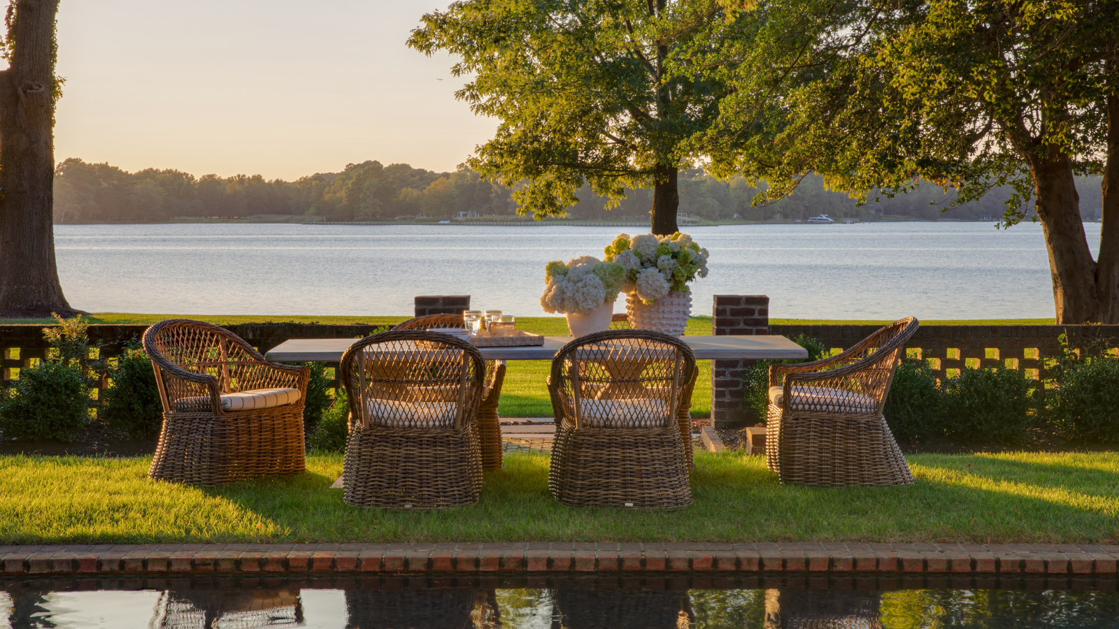 an outdoor dining table with wicker chairs around the sides on grass with a large lake in the background