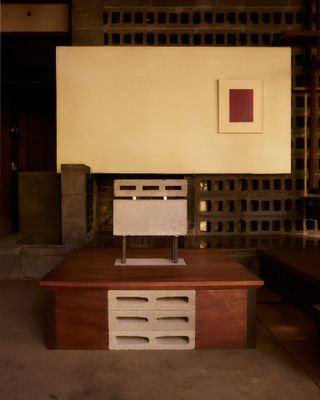Inside the Rose House, a desk with a three by tow central cutting, with a white box on top. Background wall is white with a red painting in a white border (off centre to the right).