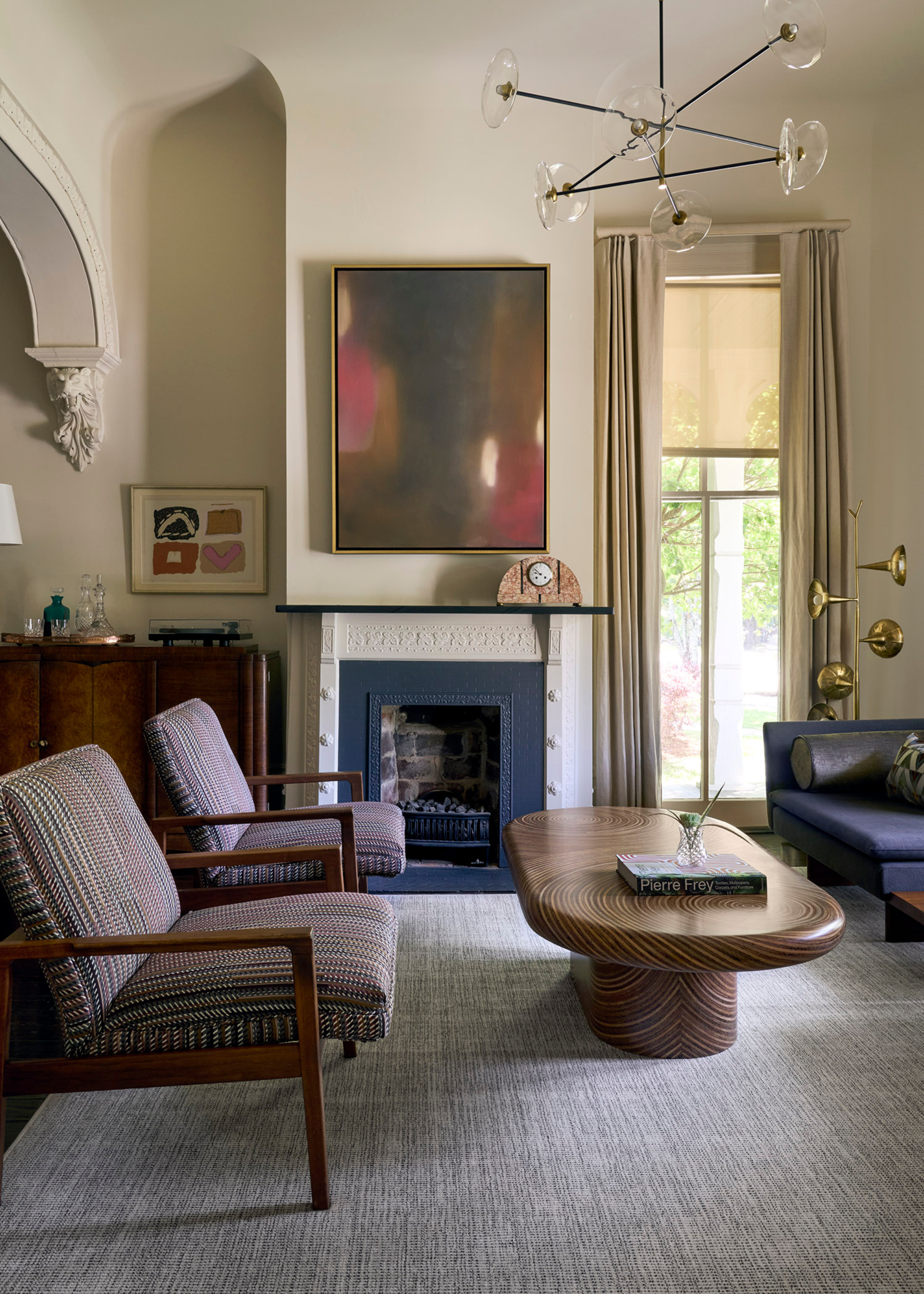 A living room with a gray area rug, patterned wood armchairs, a long wood coffee table, a blue couch, a brass floor lamp, a blue fireplace, a framed painting above and a sideboard with a bar tray and a record player beside it
