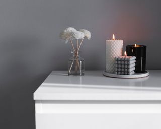 Three candles burning on a white surface next to a small vase with three white flowers in against a grey background