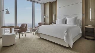 A hotel bedroom with a white bed, a desk and chair and a huge window