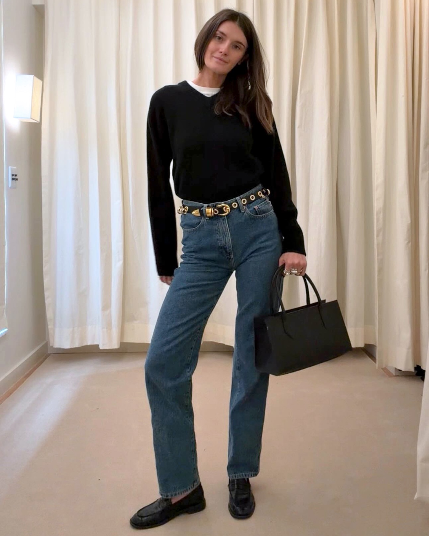 NYC based style influencer Coco Schiffer poses wearing a black V-neck sweater layered over a white tee, a black minimalist bag, studded belt, straight-leg jeans, and black loafers