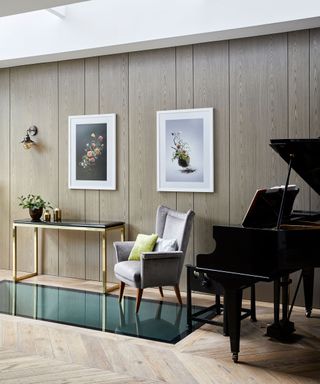 A wood panelled living room with grand piano, artwork and a gray armchair.