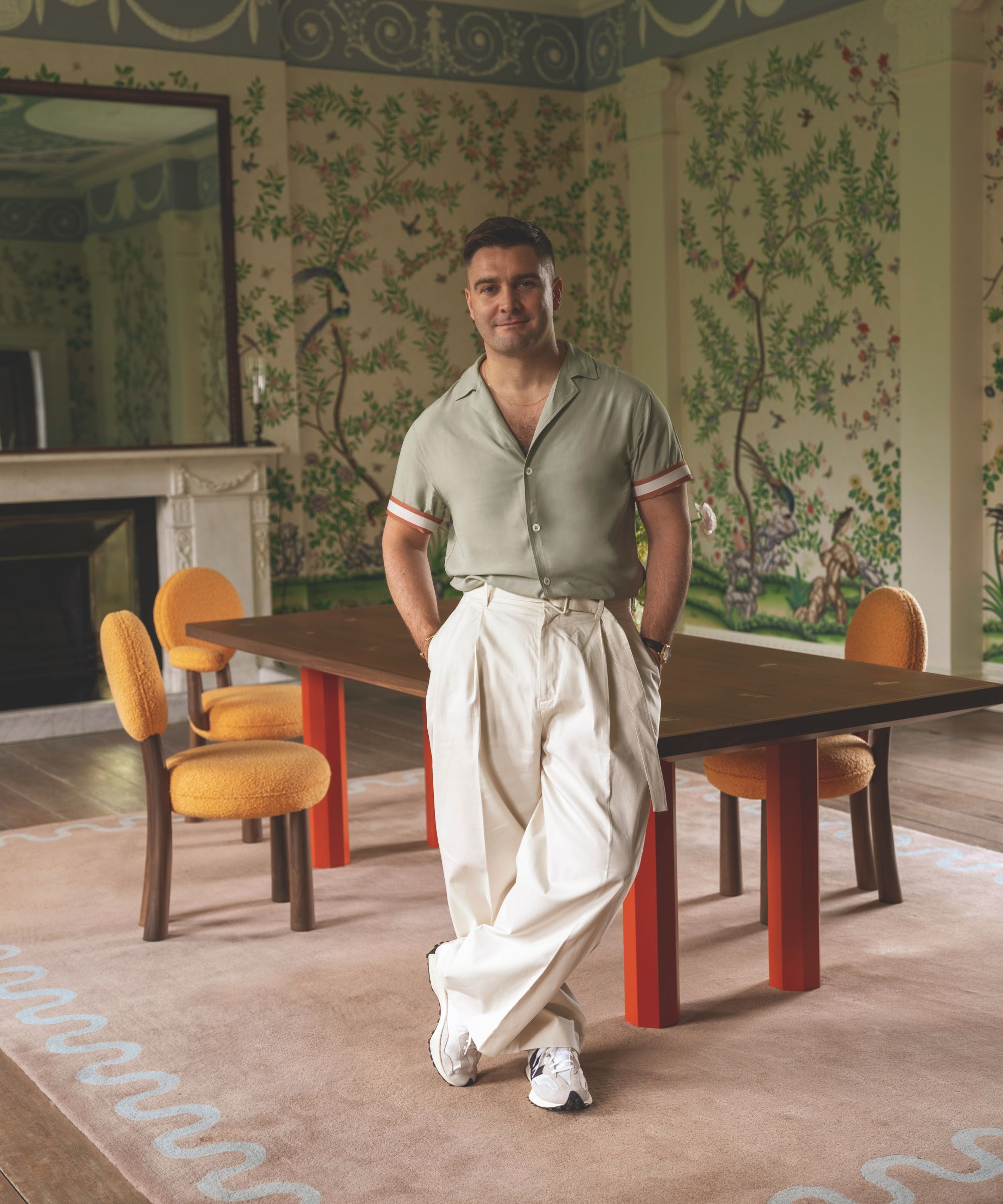A man in a grey green polo shirt and cream trousers stands in a dining room with a large pink rug and a dining table with orange chairs